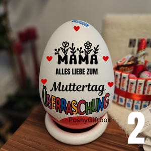 May include: A white egg-shaped gift box with the words "MAMA" and "Muttertag" in black and colorful lettering, respectively. Red hearts and floral designs decorate the egg. The base is white with a red rim.