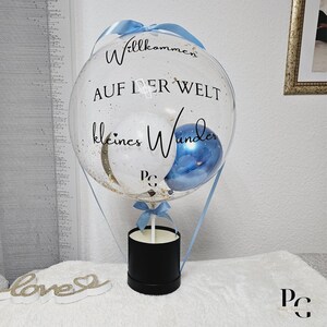 以下が含まれることがあります： 黒い文字で「Willkommen auf der Welt kleines Wunder」と書かれた、熱気球の形をした透明な風船。風船は、金色のキラキラと、白と青の小さな風船2つで満たされています。風船は黒い箱に繋がっています。