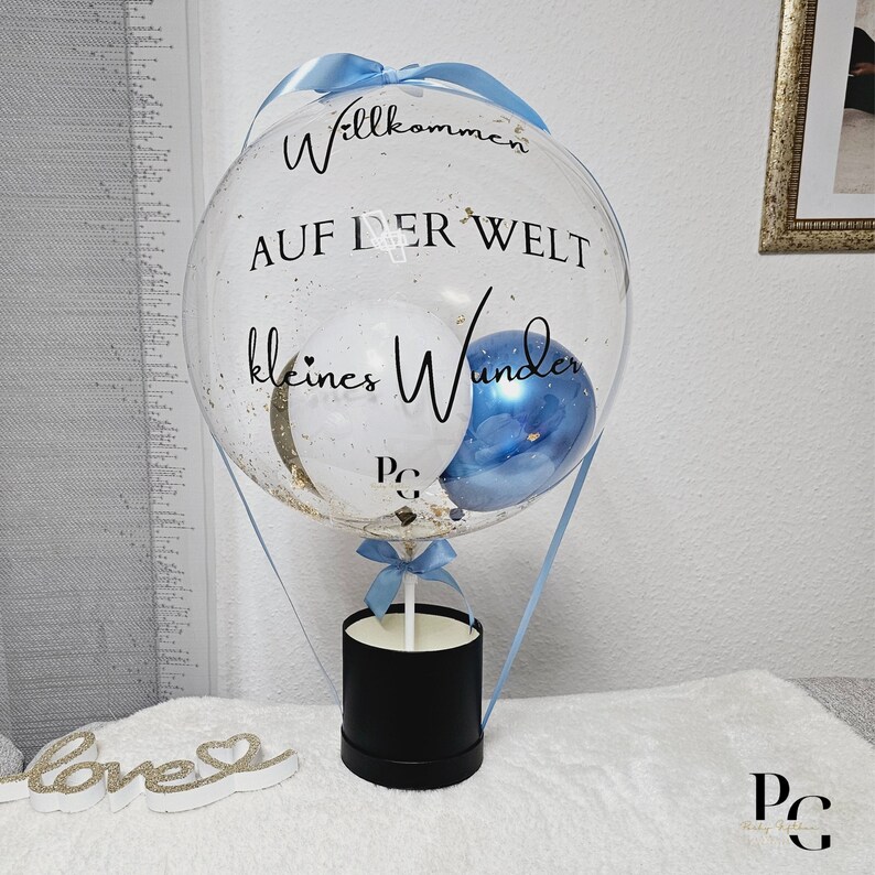 以下が含まれることがあります： 黒い文字で「Willkommen auf der Welt kleines Wunder」と書かれた、熱気球の形をした透明な風船。風船は、金色のキラキラと、白と青の小さな風船2つで満たされています。風船は黒い箱に繋がっています。