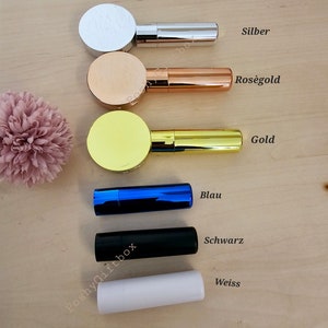 Pu&ograve; includere: Cinque tubetti di rossetto in diversi colori: argento, oro rosa, oro, blu e bianco. I tubetti sono disposti verticalmente, con il tubetto argentato in alto e il tubetto bianco in basso. Ogni tubetto ha un tappo rotondo e un'etichetta con il nome del colore in tedesco.