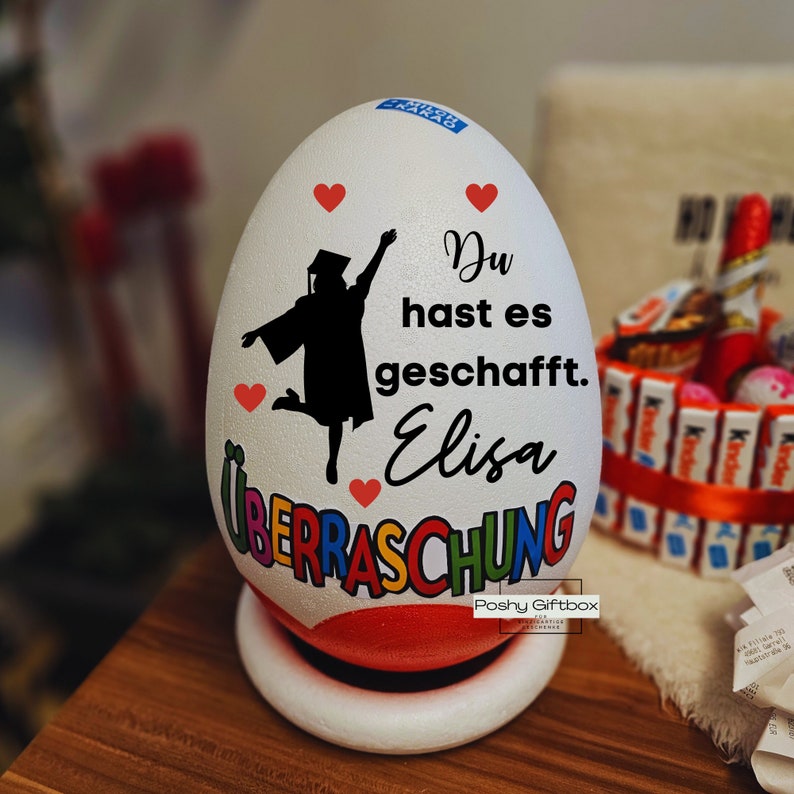 Może przedstawiać: Biały, ceramiczny jajko w kształcie jajka z sylwetką absolwenta, sercami i tekstem "Du hast es geschafft. Elisa &Uuml;BERRASCHUNG".