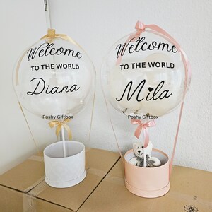 May include: Two clear balloon hot air balloons with white ribbons and a teddy bear in one of the baskets. The balloons have the text "Welcome to the world" and the names "Diana" and "Mila" on them. The balloons are sitting on top of gift boxes.