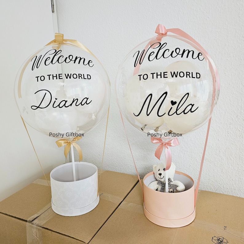 May include: Two clear balloon hot air balloons with white ribbons and a teddy bear in one of the baskets. The balloons have the text "Welcome to the world" and the names "Diana" and "Mila" on them. The balloons are sitting on top of gift boxes.