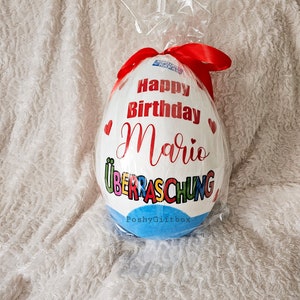 May include: A white chocolate egg wrapped in plastic with a red ribbon. The egg is decorated with the words "Happy Birthday Mario" and "&Uuml;berraschung" in colorful letters. The bottom of the egg is blue with the text "PoshyGiftbox".