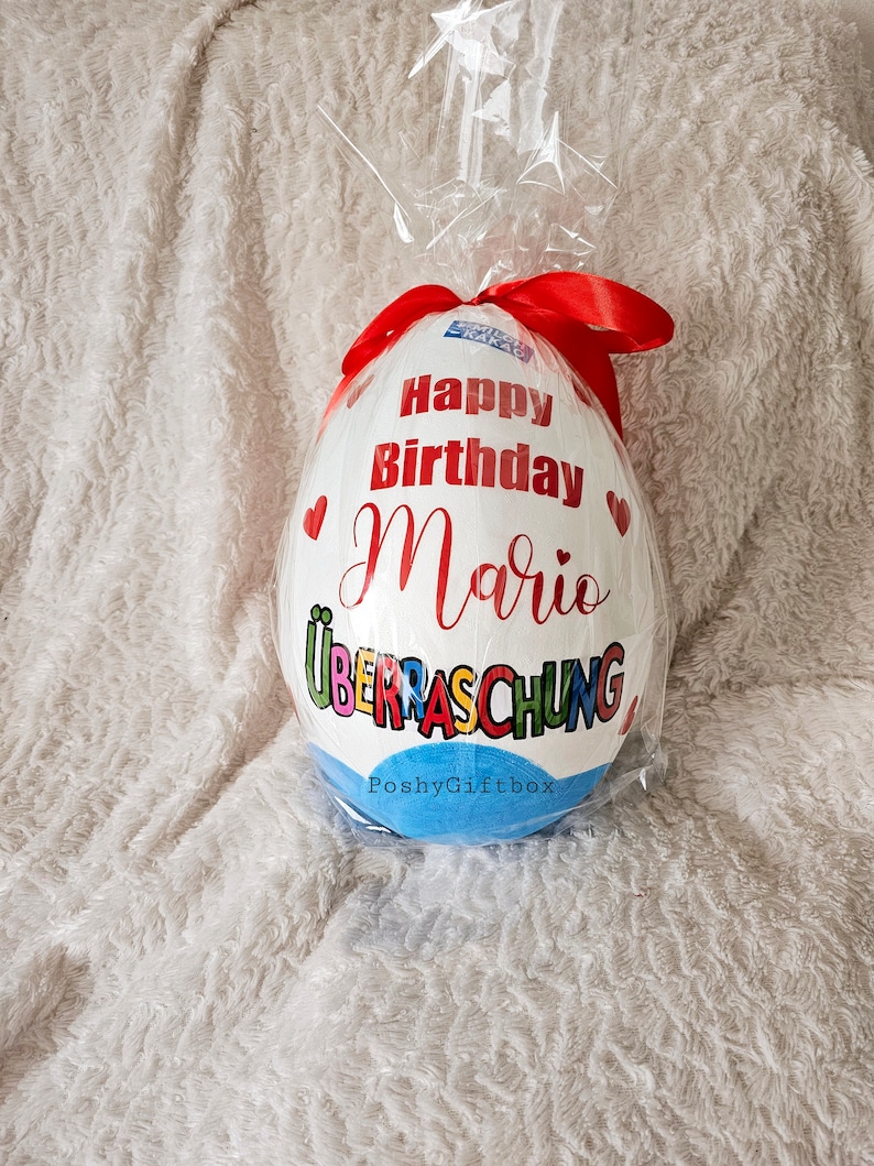 May include: A white chocolate egg wrapped in plastic with a red ribbon. The egg is decorated with the words "Happy Birthday Mario" and "&Uuml;berraschung" in colorful letters. The bottom of the egg is blue with the text "PoshyGiftbox".