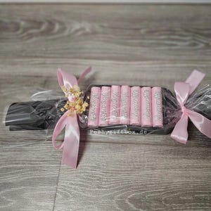 May include: A gift set containing seven pink Yogurette chocolate bars arranged in a row, wrapped in clear cellophane with black paper backing. The package is tied at both ends with light pink satin ribbons and decorated with dried flowers.