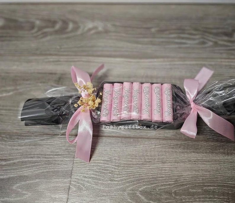 May include: A gift set containing seven pink Yogurette chocolate bars arranged in a row, wrapped in clear cellophane with black paper backing. The package is tied at both ends with light pink satin ribbons and decorated with dried flowers.