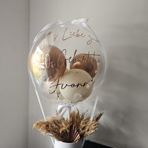 May include: A clear balloon filled with smaller metallic gold and cream balloons, featuring the rose gold text "Alles Liebe zum 40. Geburtstag Yvonne". The balloon is attached to a white cylindrical box base filled with dried pampas grass and a fan.