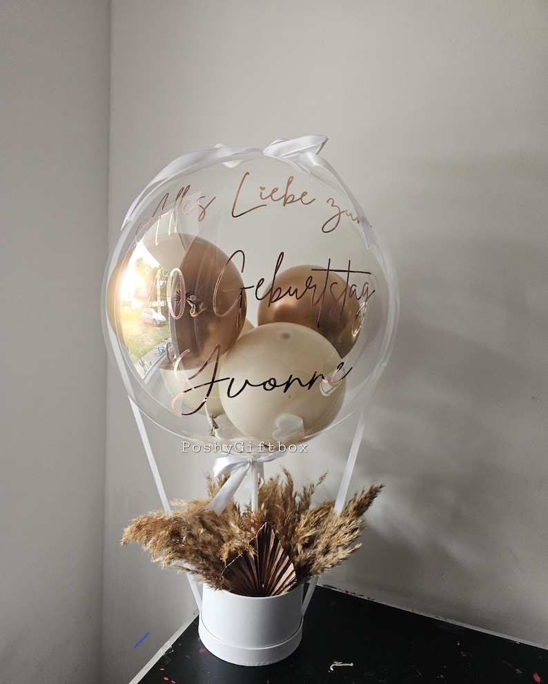 May include: A clear balloon filled with smaller metallic gold and cream balloons, featuring the rose gold text "Alles Liebe zum 40. Geburtstag Yvonne". The balloon is attached to a white cylindrical box base filled with dried pampas grass and a fan.