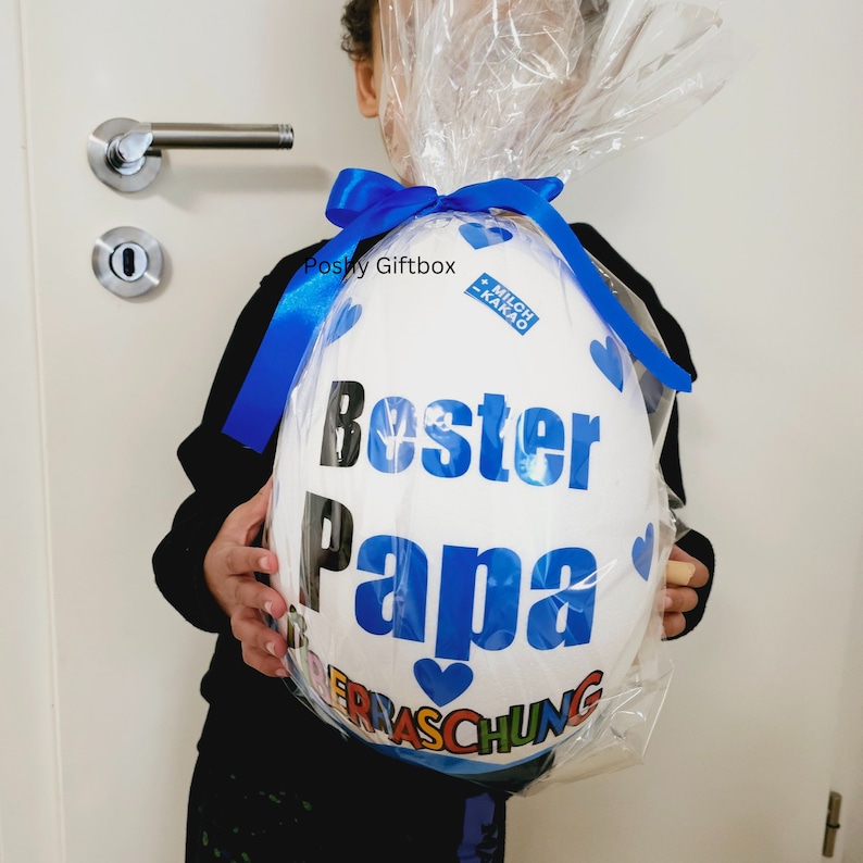 以下が含まれることがあります： 青いリボンと「Bester Papa」という文字が書かれた、大きな白い卵型のギフトボックス。ボックスは透明なプラスチックで包まれ、小さな青いハートのステッカーが貼られています。