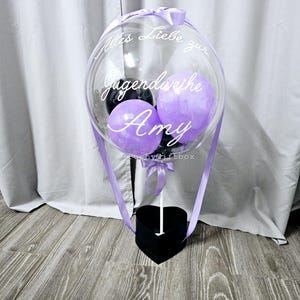 Op de afbeelding: Een doorzichtige, ronde ballon met kleinere paarse en zwarte ballonnen erin, bevestigd op een zwarte hartvormige doos. De ballon is voorzien van de witte cursieve tekst "Alles Liebe zur Jugendweihe Amy" en een lichtpaars lint.