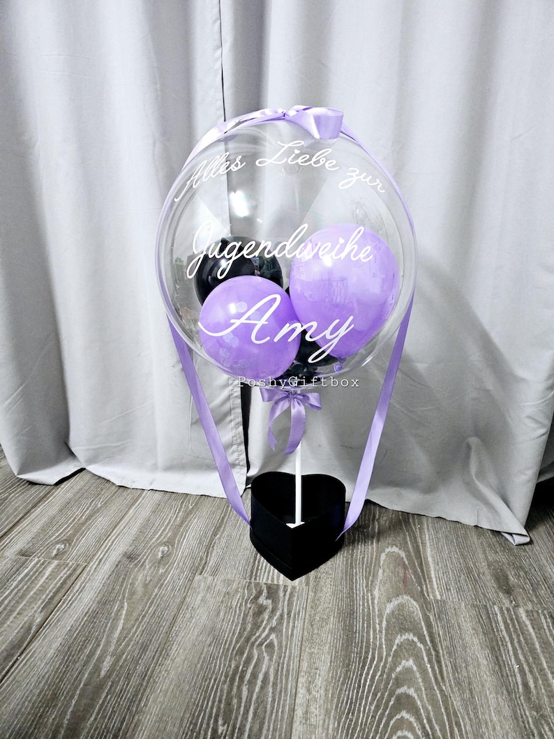 Op de afbeelding: Een doorzichtige, ronde ballon met kleinere paarse en zwarte ballonnen erin, bevestigd op een zwarte hartvormige doos. De ballon is voorzien van de witte cursieve tekst "Alles Liebe zur Jugendweihe Amy" en een lichtpaars lint.