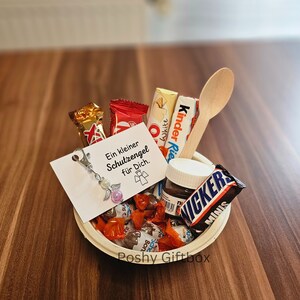 May include: A white paper bowl filled with various chocolate candies, a small jar of chocolate spread, a wooden spoon, and a small white card with the text "Ein kleiner Schutzengel f&uuml;r Dich."  The bowl is labelled "Poshy Giftbox".