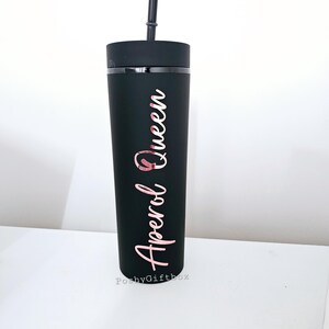May include: Black tumbler with a rose gold inscription that reads "Aperol Queen". The tumbler has a black lid and a black straw.