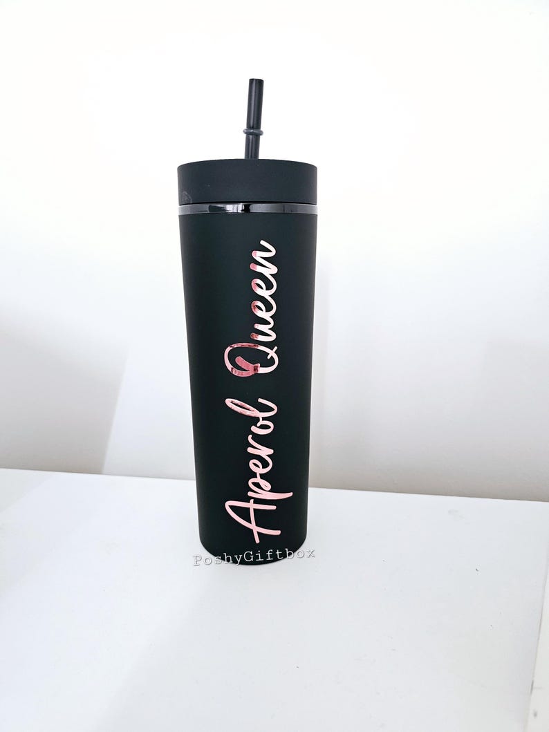 May include: Black tumbler with a rose gold inscription that reads "Aperol Queen". The tumbler has a black lid and a black straw.