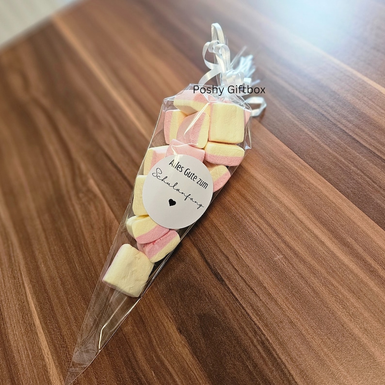 May include: A clear plastic cone filled with pink and yellow marshmallows. A white circle sticker with the text "Alles Gute zum Schulanfang" and a small heart is attached to the cone.