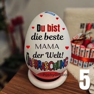 May include: A white egg-shaped gift box with red heart accents and the German text "Du bist die beste MAMA der Welt!" in black and red. The word "&Uuml;BERRASCHUNG" is in colorful letters. The gift box sits on a white and red base.
