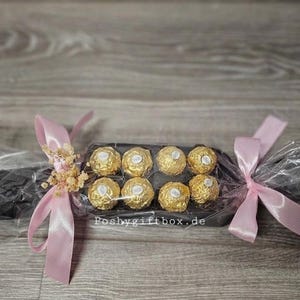 May include: A black rectangular gift box containing eight gold-wrapped spherical chocolates, wrapped in clear cellophane with light pink satin ribbons tied at both ends and small dried flowers, with the text poshygiftbox.de printed on the centre.