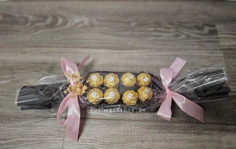 May include: A black rectangular gift box containing eight gold-wrapped spherical chocolates, wrapped in clear cellophane with light pink satin ribbons tied at both ends and small dried flowers, with the text poshygiftbox.de printed on the centre.
