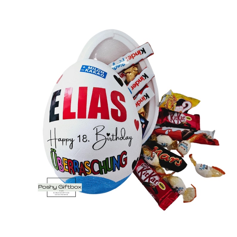 Może przedstawiać: Duże, białe pudełko w kształcie jaja z napisem "ELIAS" w kolorze czerwonym i "Happy 18. Birthday". Pudełko jest wypełnione r&oacute;żnymi batonami czekoladowymi, w tym Kinder i KitKat. Pudełko jest otwarte, a niekt&oacute;re cukierki wypadają.