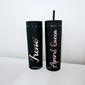 May include: Two black reusable tumblers with lids and straws. The tumbler on the left has the name "Irene" written in gold lettering. The tumbler on the right has the text "Aperol Queen" written in gold lettering.