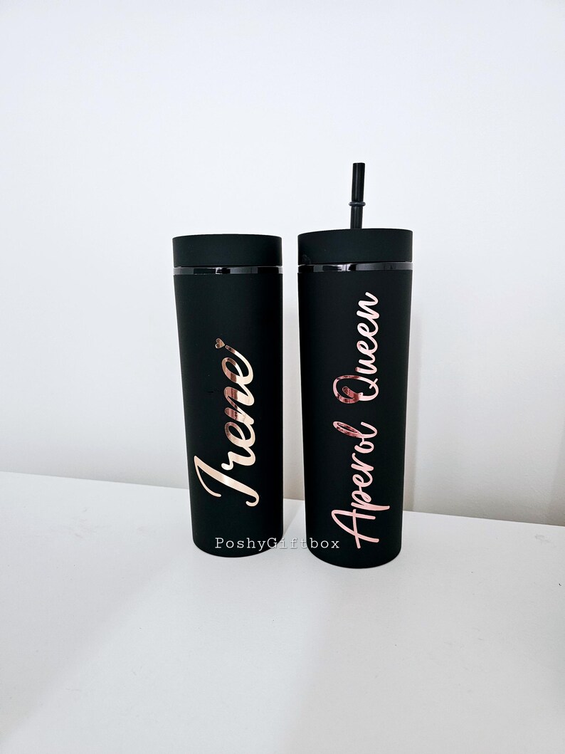 May include: Two black reusable tumblers with lids and straws. The tumbler on the left has the name "Irene" written in gold lettering. The tumbler on the right has the text "Aperol Queen" written in gold lettering.