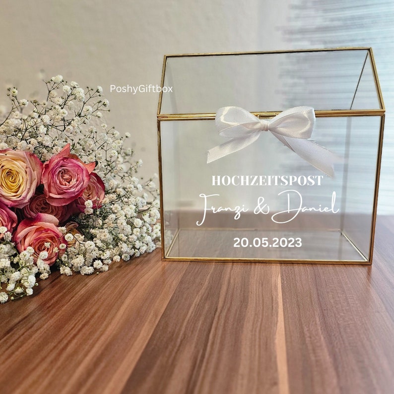 May include: A clear glass box with a gold frame and a white bow on top. The box has the text "HOCHZEITSPOST Franzi & Daniel 20.05.2023" printed on the front.