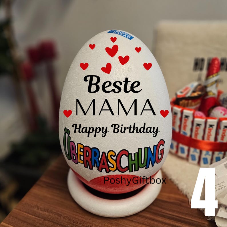 May include: A white egg-shaped gift box with red heart accents and the words "Beste MAMA Happy Birthday UBERRASCHUNG" in black and colorful lettering. The gift box sits on a white base with a red rim. The words "PoshyGiftbox" are also visible.