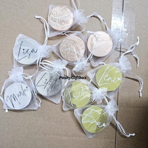 Pu&ograve; includere: Una collezione di sottobicchieri rotondi in legno personalizzati con nomi scritti in corsivo. I sottobicchieri sono confezionati in sacchetti individuali di organza bianca con nastri bianchi. I sottobicchieri sono esposti su una superficie marrone.