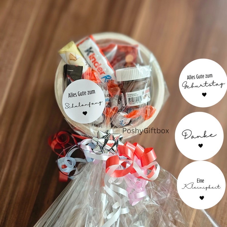 May include: A white gift basket filled with various treats, including chocolate bars, candy, and a jar of Nutella. The basket is wrapped in clear cellophane and tied with red, white, and pink ribbons. The basket has a white tag that says "Alles Gute zum Schulanfang" and a small red heart. There are three other white tags with black text that say "Alles Gute zum Geburtstag", "Danke", and "Eine Kleinigkeit".