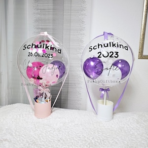 May include: Two clear balloon hot air balloons with purple and pink confetti inside. The balloons are decorated with the text "Schulkind 2023" and "Schulkind 26.06.2023" with names "Sofia" and "Chaya Jadyn". The balloons are attached to white boxes with purple and pink ribbons.