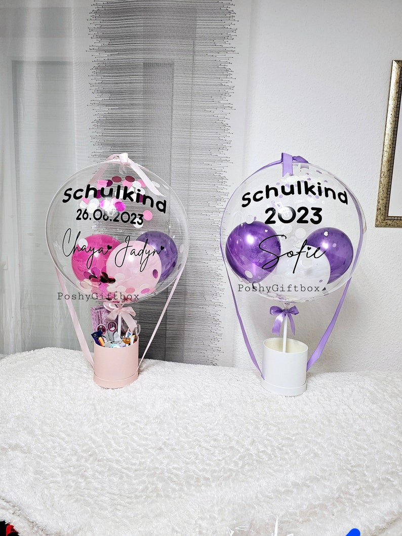May include: Two clear balloon hot air balloons with purple and pink confetti inside. The balloons are decorated with the text "Schulkind 2023" and "Schulkind 26.06.2023" with names "Sofia" and "Chaya Jadyn". The balloons are attached to white boxes with purple and pink ribbons.