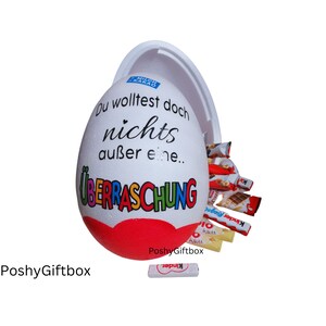 May include: A white plastic egg with a red heart and the text "Du wolltest doch nichts au&szlig;er eine... &Uuml;BERRASCHUNG" in rainbow colors. The egg is open and filled with various chocolate bars and candies.
