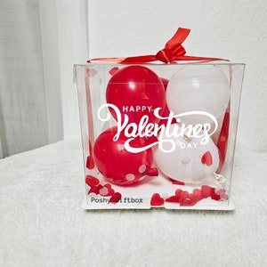 May include: A clear plastic box filled with red and white balloons. The balloons have the text "Happy Valentines Day" printed on them. The box is decorated with small red hearts.