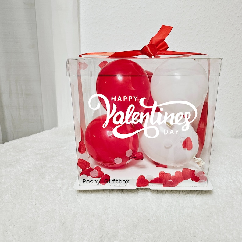 May include: A clear plastic box filled with red and white balloons. The balloons have the text "Happy Valentines Day" printed on them. The box is decorated with small red hearts.