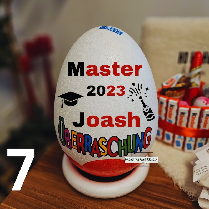 Może przedstawiać: Biały pojemnik w kształcie jajka z napisem "Master 2023 Joash Uberraschung" w czarnych i czerwonych literach. Na pojemniku przedstawiono czapkę absolwenta i butelkę szampana. Pojemnik stoi na białej podstawie.