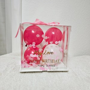 May include: A clear plastic box filled with pink and white balloons and pink confetti. The box has a pink ribbon and a gold sticker that says "My Love Happy Birthday Poshy Giftbox".
