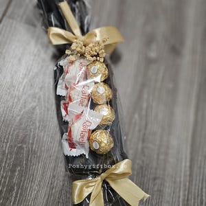 May include: A gift arrangement wrapped in clear cellophane featuring a black base, four gold-foiled spherical chocolates, and three white-wrapped Raffaello candies. The bundle is decorated with dried flowers and two gold satin ribbons, with Poshygiftbox.de printed on the front.