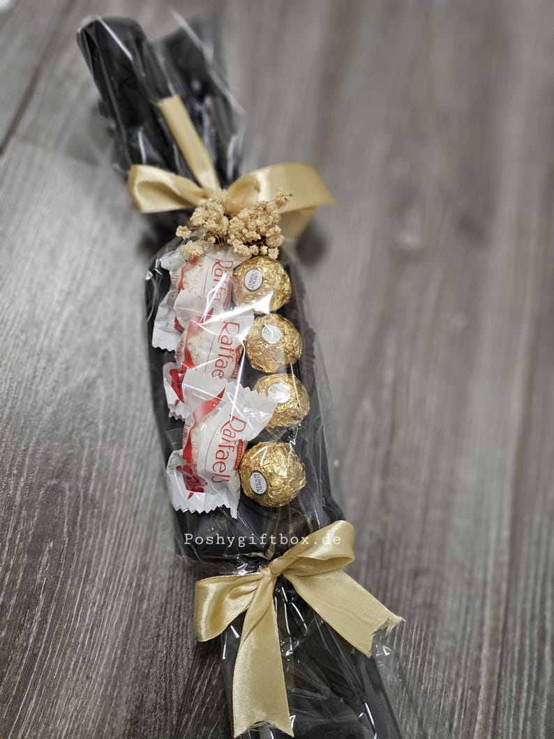 May include: A gift arrangement wrapped in clear cellophane featuring a black base, four gold-foiled spherical chocolates, and three white-wrapped Raffaello candies. The bundle is decorated with dried flowers and two gold satin ribbons, with Poshygiftbox.de printed on the front.