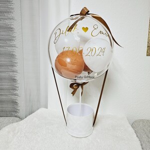 May include: A clear balloon containing smaller white and brown balloons, attached to a white cylindrical base with brown ribbons. The clear balloon features gold text reading Dilek, a heart symbol, Emre, and the date 17.05.2024, with Poshy Giftbox printed below.