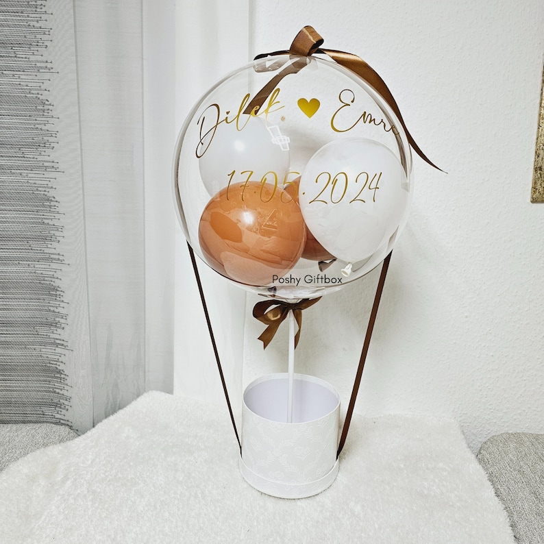 May include: A clear balloon containing smaller white and brown balloons, attached to a white cylindrical base with brown ribbons. The clear balloon features gold text reading Dilek, a heart symbol, Emre, and the date 17.05.2024, with Poshy Giftbox printed below.