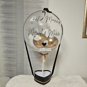 May include: A clear balloon hot air balloon with a black ribbon, two gold balloons, and white paper hearts inside. The balloon has the text "Just Married Marcel & Mari" written on it. The balloon is attached to a black heart-shaped box with a gold ribbon.