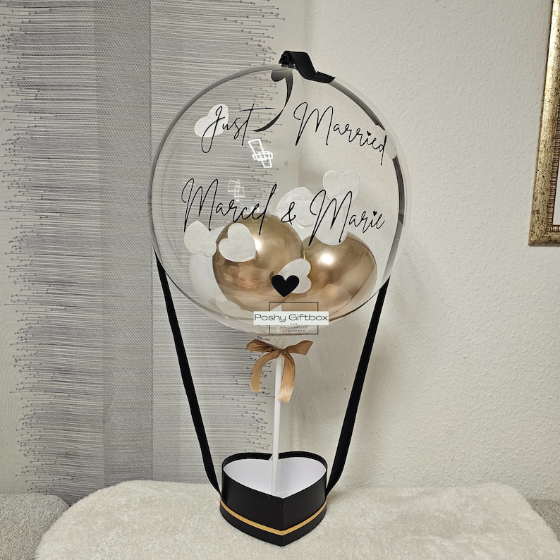 May include: A clear balloon hot air balloon with a black ribbon, two gold balloons, and white paper hearts inside. The balloon has the text "Just Married Marcel & Mari" written on it. The balloon is attached to a black heart-shaped box with a gold ribbon.
