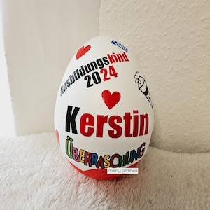 Może przedstawiać: Biały pojemnik w kształcie jajka z czerwonym, czarnym i niebieskim tekstem i sercami. Tekst brzmi "Ausbildunkskind 2024" i "Kerstin" z mniejszym tekstem, kt&oacute;ry brzmi "&Uuml;berraschung" i "Poshy Giftbox".