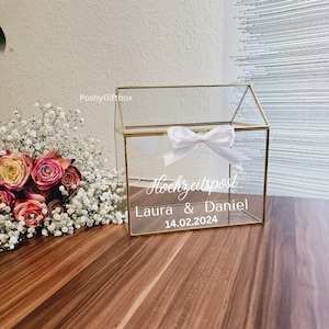 May include: A clear glass house-shaped box with a gold trim and a white ribbon bow. The box has a sign that says "Hochzeitspost Laura & Daniel 14.02.2024".
