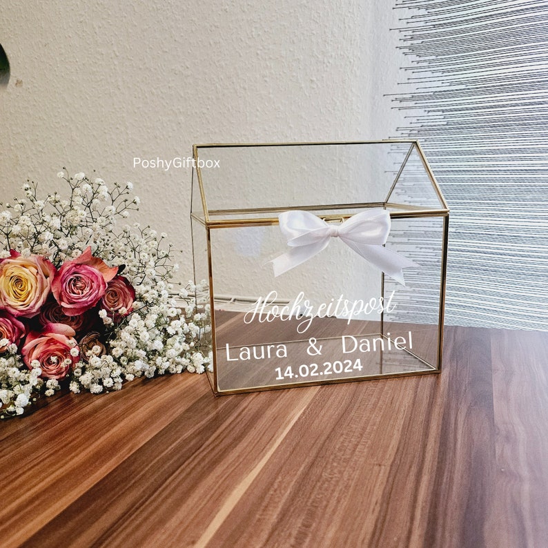 May include: A clear glass house-shaped box with a gold trim and a white ribbon bow. The box has a sign that says "Hochzeitspost Laura & Daniel 14.02.2024".