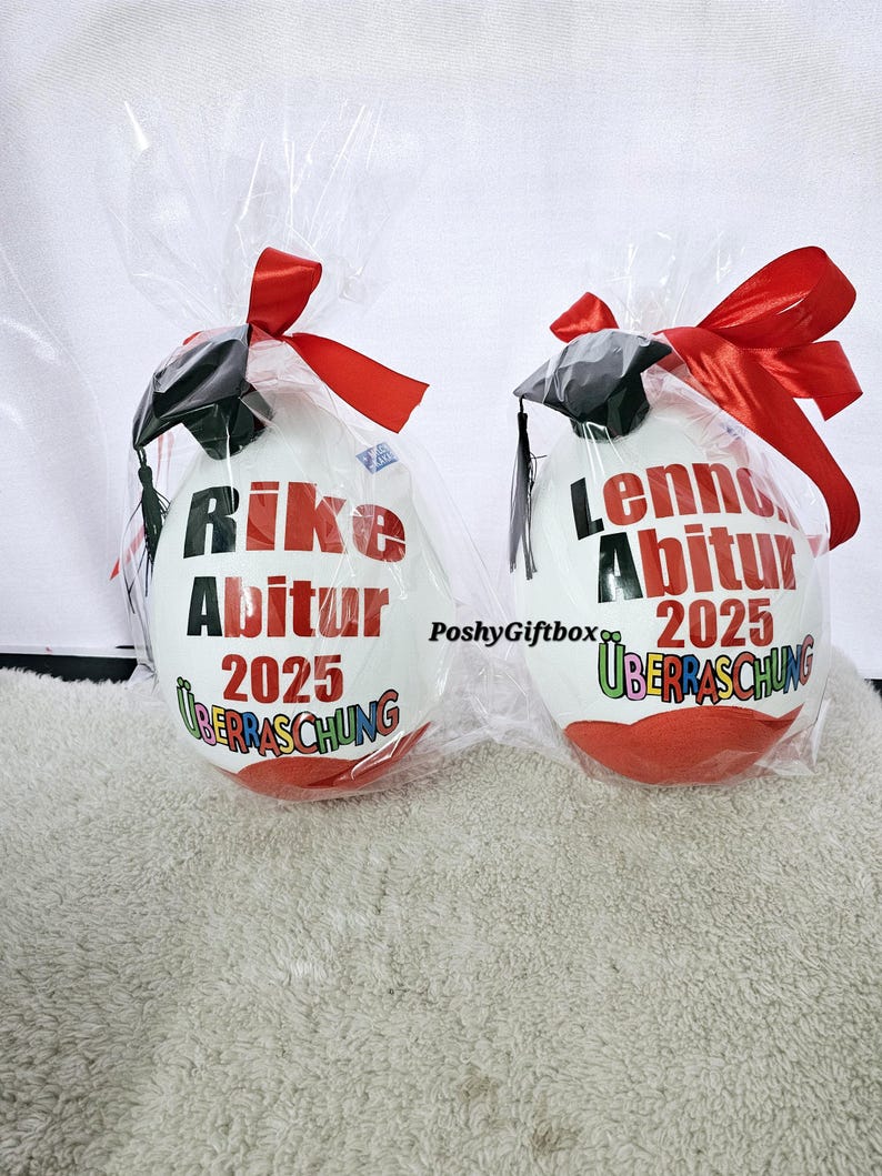 May include: Two white graduation balloons with red, black, and colorful accents. The balloons are decorated with the text "Rike Abitur 2025" and "Lenn Abitur 2025" with the word "&Uuml;berraschung" underneath each name.