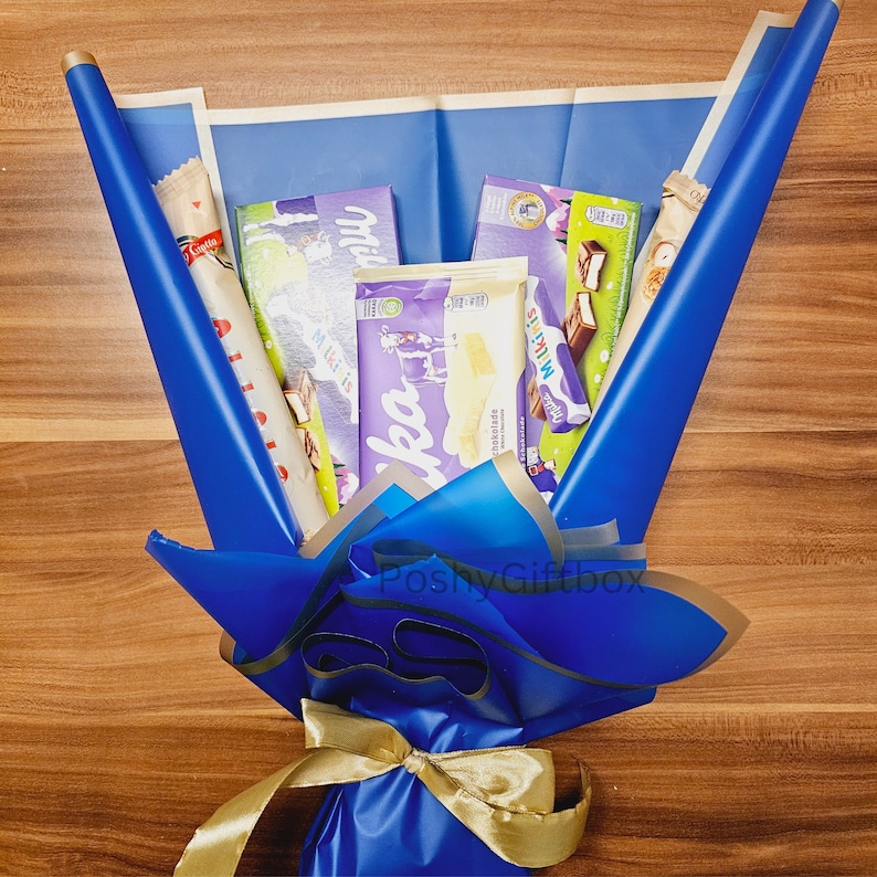 May include: A blue gift bouquet wrapped in blue tissue paper with a gold ribbon. The bouquet contains several chocolate bars, including Milka chocolate bars.