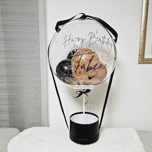 May include: A clear balloon with black and gold confetti inside, tied to a black ribbon and a black box. The balloon has the text "Happy Birthday" and "Tabea" written on it. The box has a white top and a black base. The balloon is attached to a stick with a black ribbon and a tag that says "Poshy Giftbox".