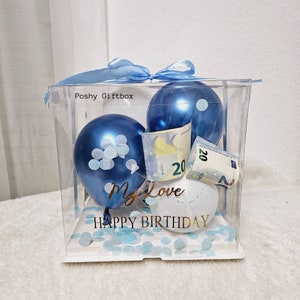 May include: A clear plastic gift box with a blue ribbon, two blue balloons, a white balloon, blue confetti, and a twenty euro bill. The box has a white base and the text "Poshy Giftbox" and "My Love Happy Birthday" printed on it.